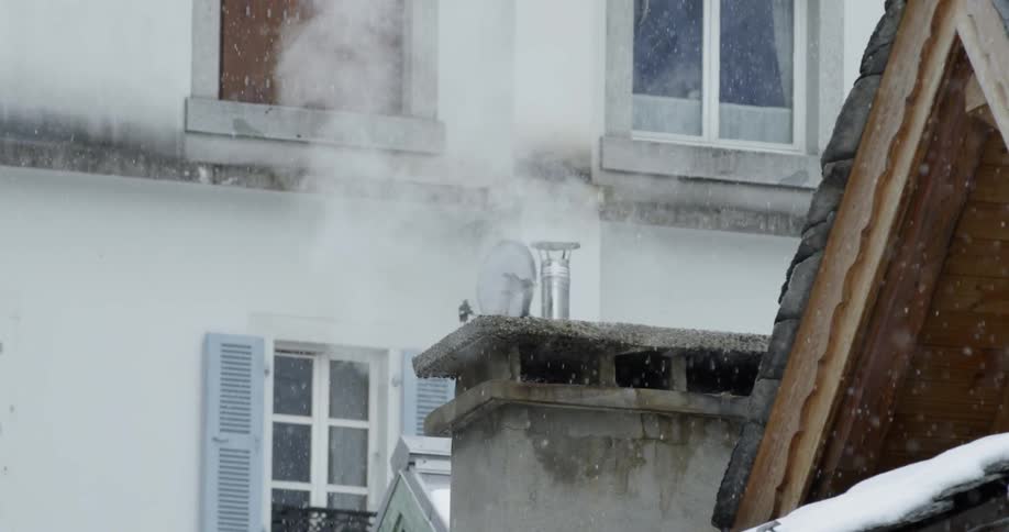 The snow is falling as steam rises from a building chimney