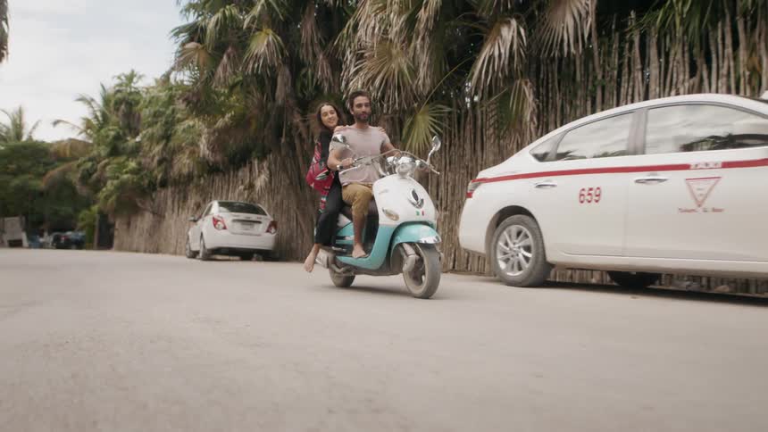 A couple are riding a moped on a road in a tropical setting during the day