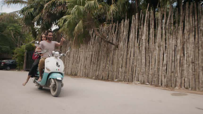Man and woman ride a small motorbike past a wooden fence