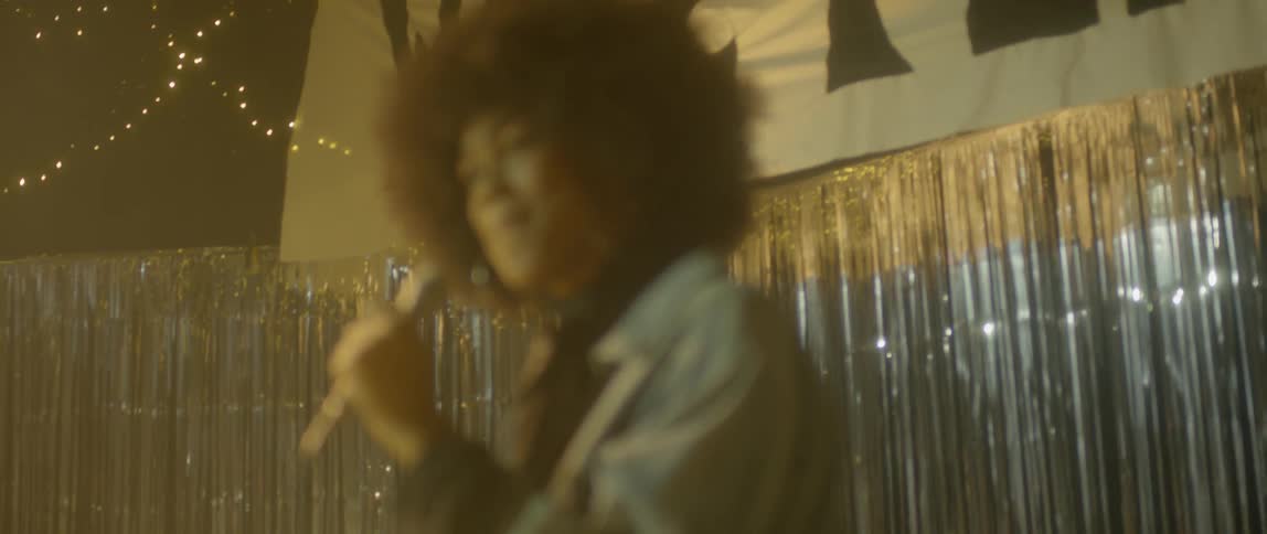 A woman sings into a microphone with lights and banner behind her