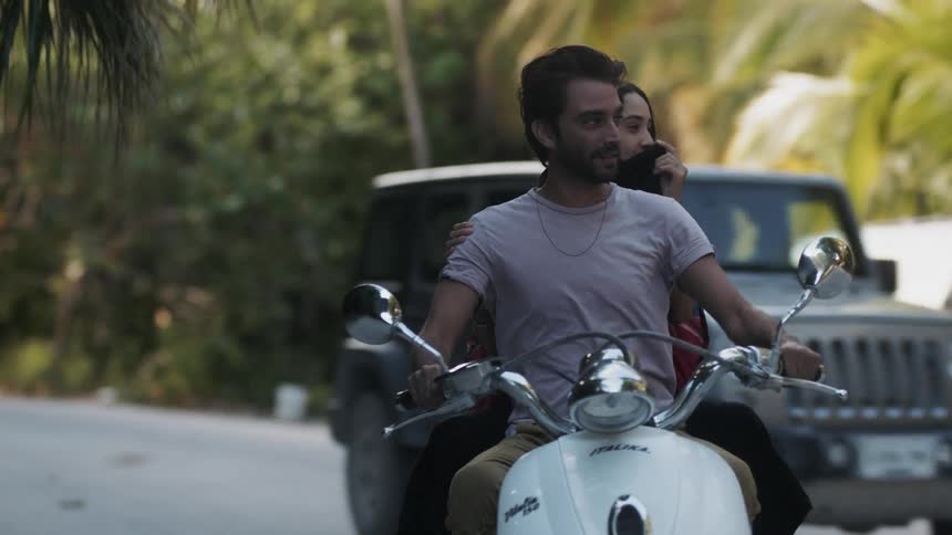 A couple are riding a moped on a road in a tropical setting during the day