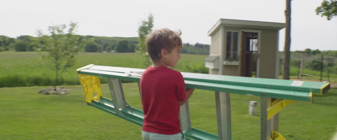 A boy carrying a ladder across his yard