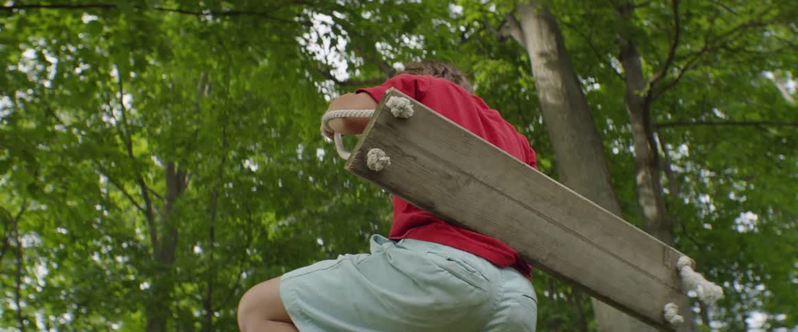 A boy is climbing up into a tree swing during the day