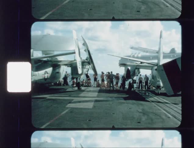 Military aircraft sit on a ship as onlookers look on