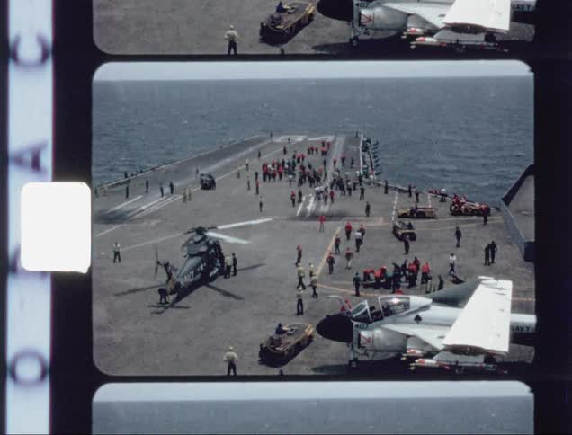 People are milling about on an aircraft carrier deck as a helicopter prepares for flight