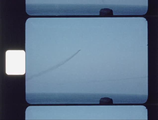 Men standing on a ship's deck are watching a fighter jet fly through the sky