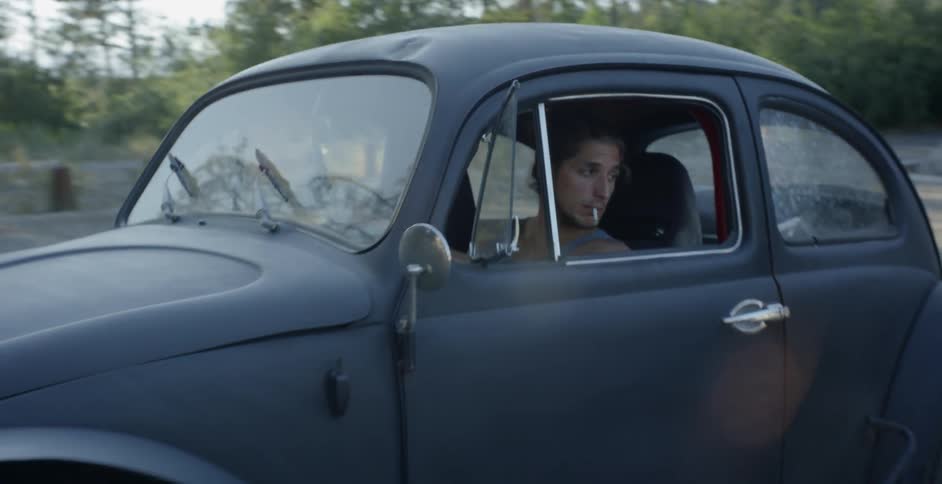 A man is backing an old model vw bug out of a driveway during the day