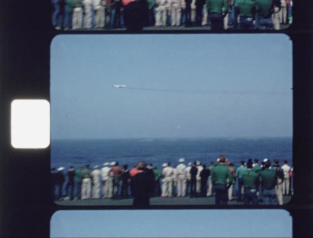 A jet flies over the water dropping bombs as a group of people watch from afar