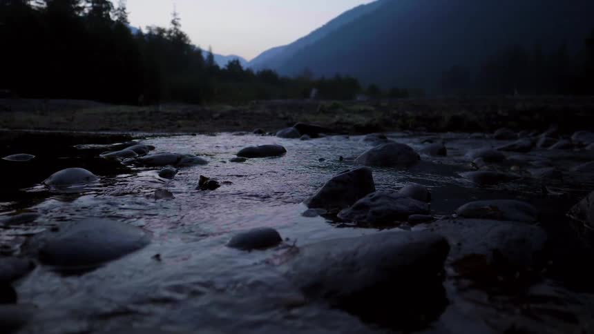 Water flowing through large rocks with a beach, tall trees and mountains in the background