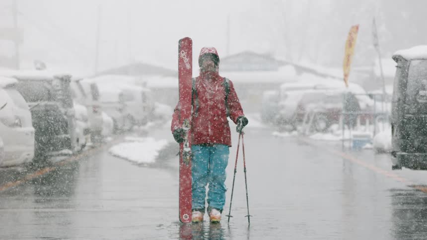 A skier stands in a mountain resort parking lot while it snows