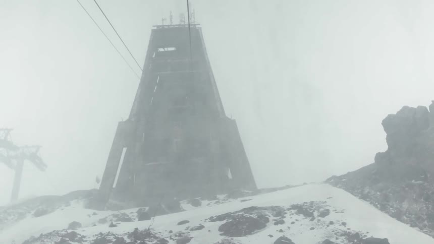 Snow flurries are blowing across a ski lift tower and mountainside during the day
