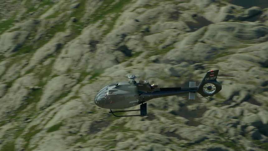 A helicopter flies above a lake and stone mountains