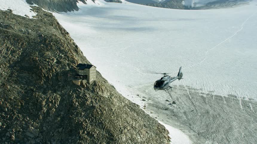 A helicopter is flying above a building in a mountain range in daylight