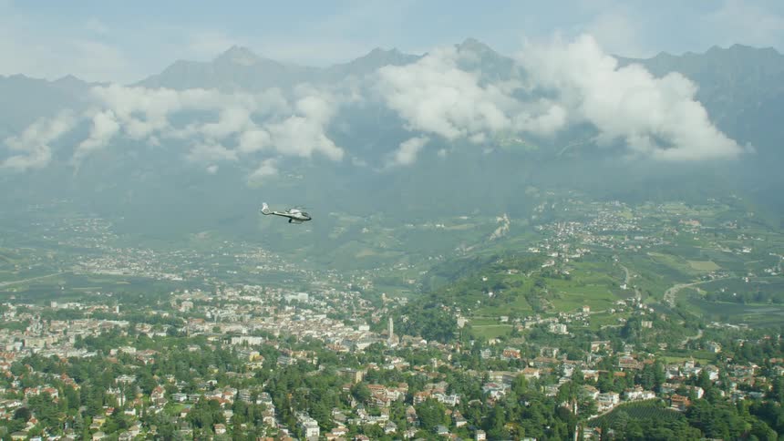 A helicopter flying over a town with many trees and buildings in front of clouds and mountains