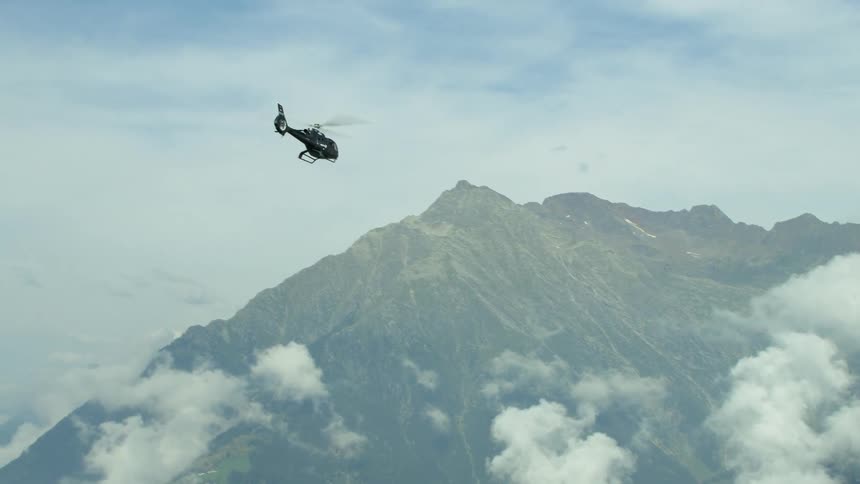 A helicopter flying backwards over mountains with clouds in the sky