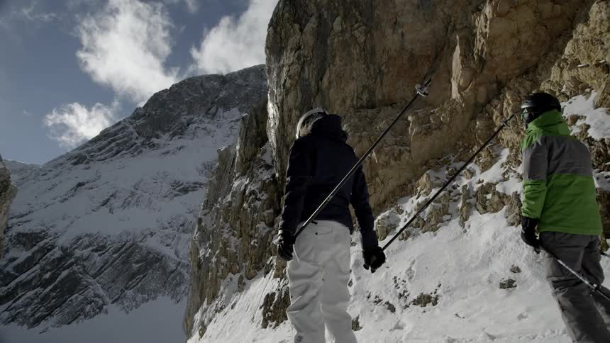 Two skiers travel down a slight slope between tall mountains