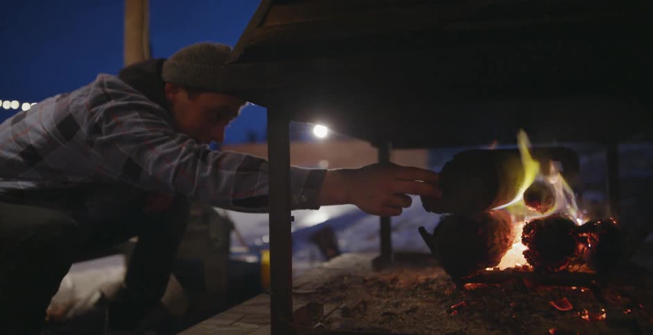 A man is putting a log onto a fire in an outdoor chimney at night
