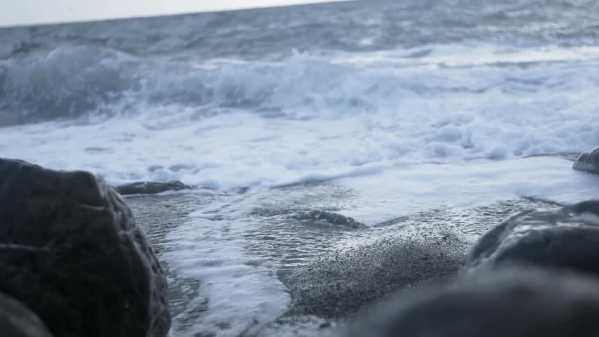 A wave is rolling toward rocks on a beach during the day