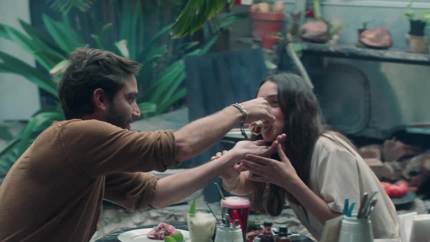 A man is offering a woman a bite of his food as they sit outside in a restaurant