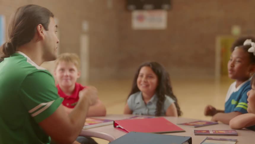 A teacher is interacting with students sitting at a table in a school building in daylight