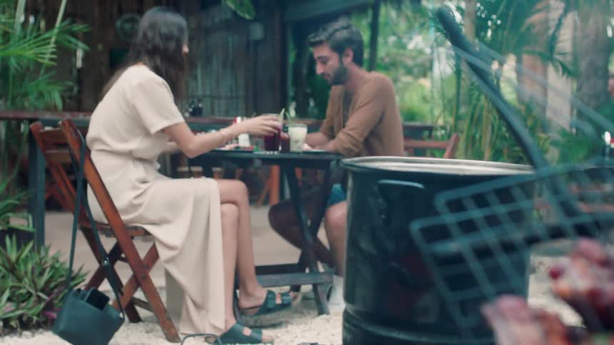 A couple sits at an outdoor restaurant as food is grilled nearby