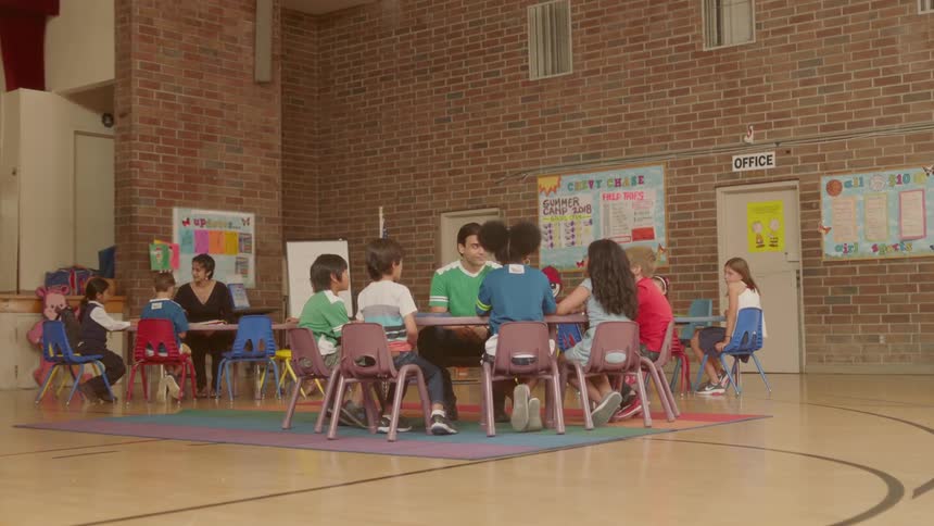 Two teachers sit at large tables with young school children in a gymnasium