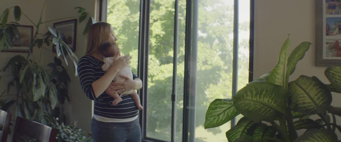 Woman holding an infant next to an open window while patting its back on a windy day