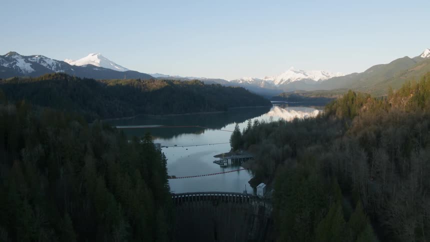 Pullback reveal of a hydroelectric dam surrounded by dense woods and snow capped mountains in the distance