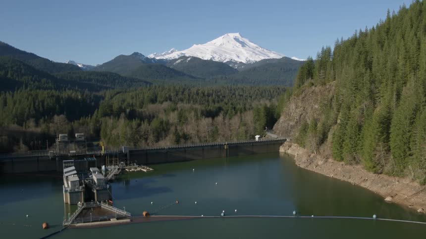 A wide concrete dam holds back water near a forest and mountain