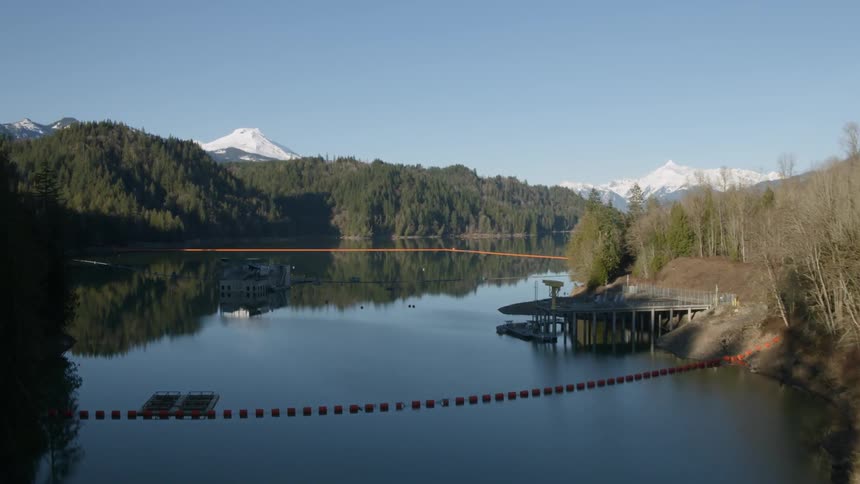 Flying just above the rim of a tall dam to reveal a deep reservoir surrounded by forest and mountains