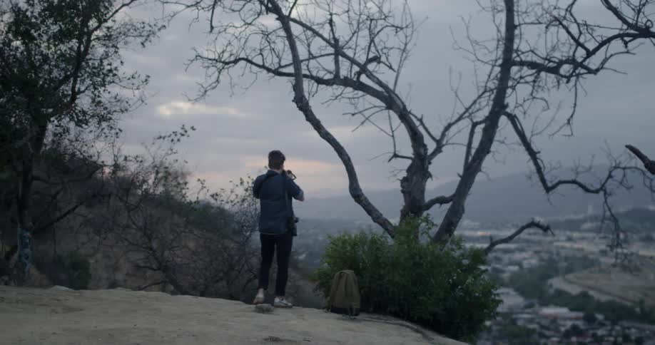 Man walks outside by bushes and trees and takes a picture of the water below