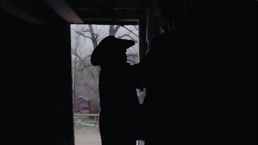 A cowboy carries a saddle out of the barn