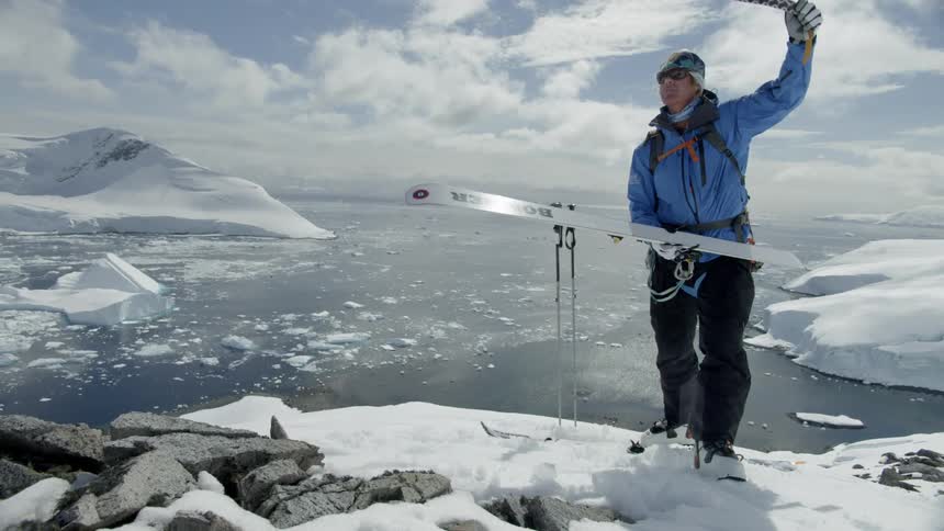 Man standing on snowy rocky ground near a body of water rips tape off of his ski