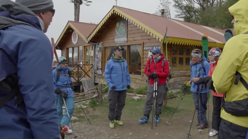 Group of people holding ski poles talk outside of a brown cabin