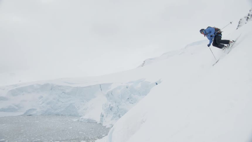 Man skis down a snowy mountain near a lake while wearing a backpack