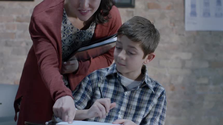 Child writes at a desk and a woman points to something on his paper