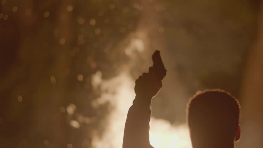 A man stands in front of a fire at night holding a gun in the air