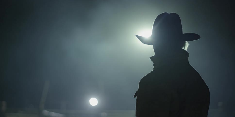 A tractor loader drives past a cowboy standing outside at night