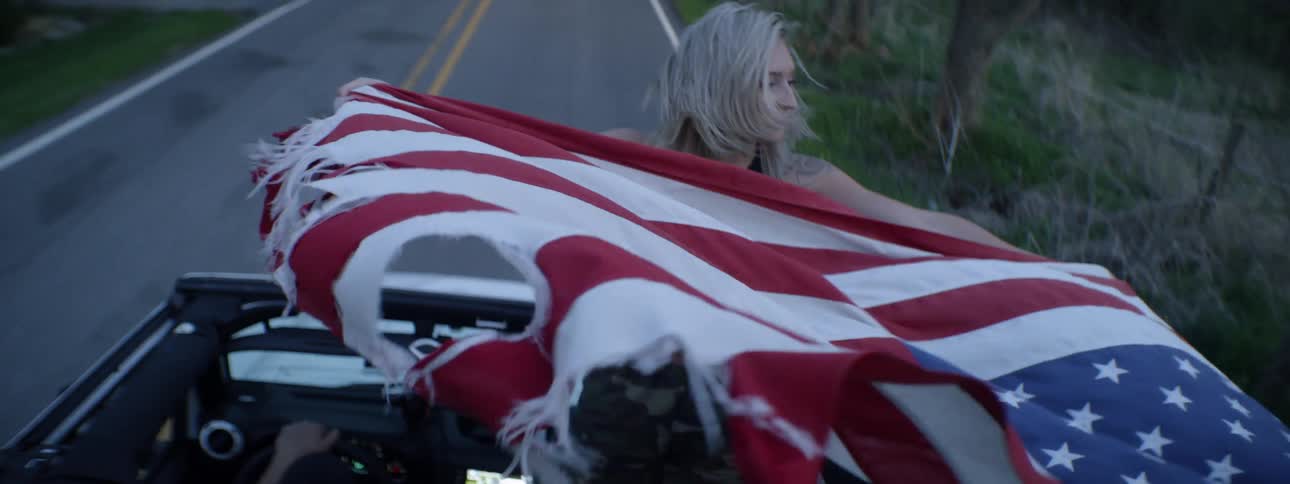 A woman stands in a convertible car and waves an American flag