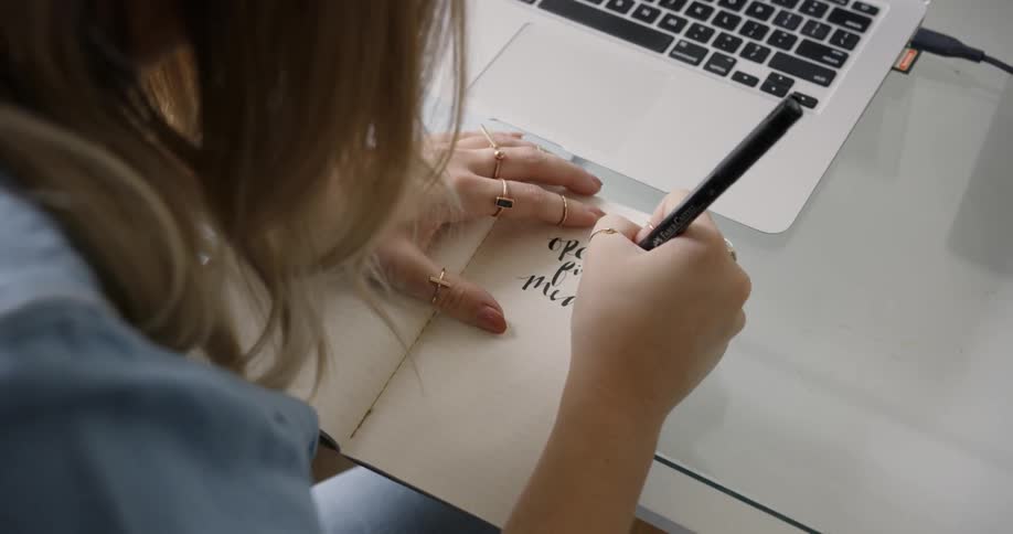 Female College Student Artist writing in a journal on a table with a laptop in front of her