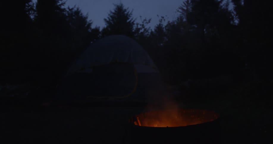 A campfire burns outside of a tent at night