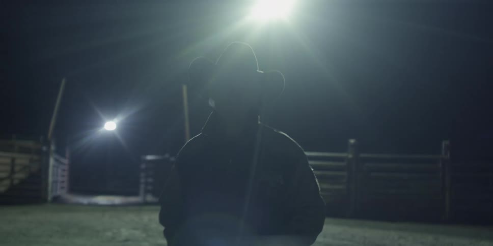 Man wearing a hat walks through a dirt yard surrounded by a fence at night