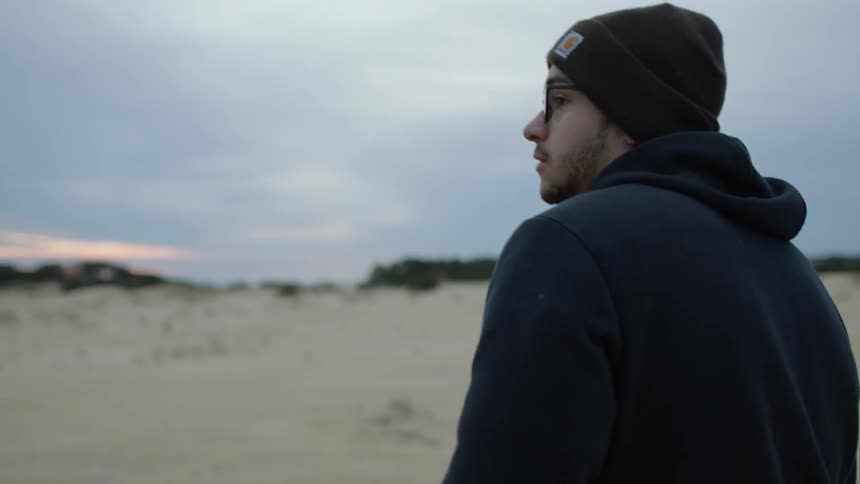 A man wearing a hat walks in the sand at sunset