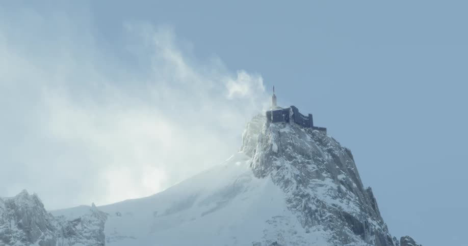 Wind blows snow off a mountain peak in europe with building perched on top