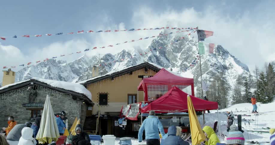 People gathered outside a mountain chalet on a windy day as flags blow