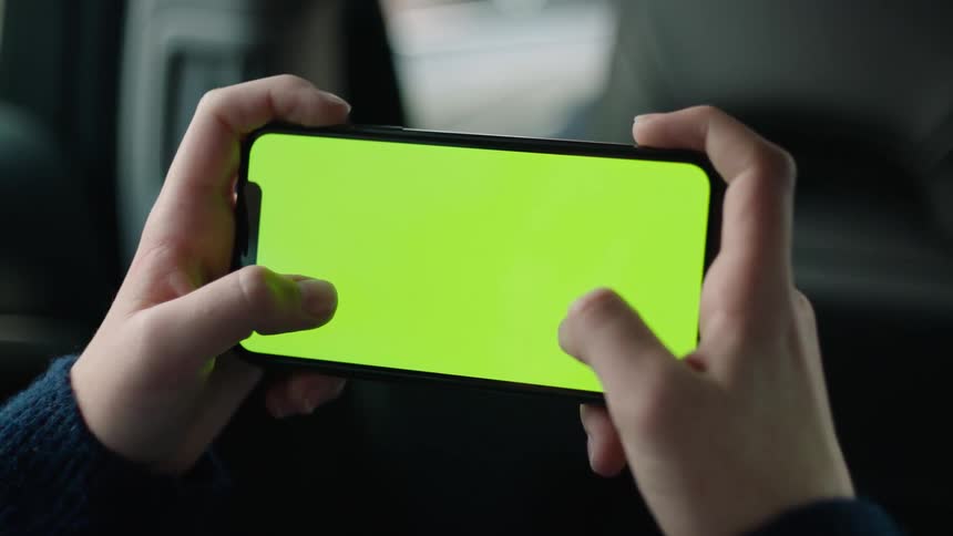 A cinematic close-up of hands holding a smartphone with a green screen and tracking markers in landscape mode inside a car during the day. The person lowers the phone at the end of the shot, perfect for VFX, screen replacement, and app mockups.
