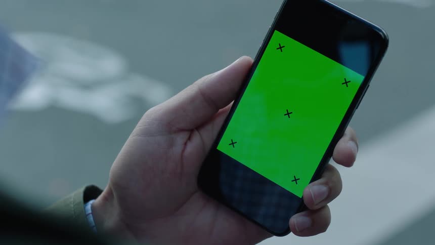 Close-up of a person standing by a street holding a smartphone with a green screen and tracking markers in daylight. The phone is held steady without interaction, perfect for VFX, screen replacement, compositing, and mobile app or tech projects.