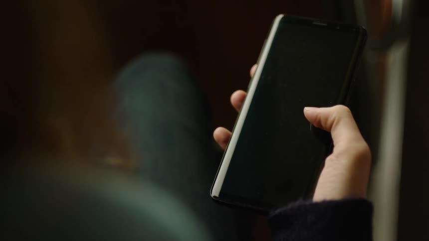 Close-up of a person sitting on a bus holding a smartphone with a black screen and moving their thumb across the display. Filmed in soft natural light, ideal for VFX, screen replacement, app mockups, UI design, and modern commuting scenes.