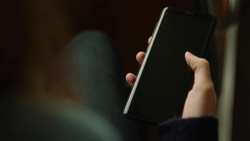 Close-up of a person sitting on a bus holding a smartphone with a black screen and repeatedly swiping up with their thumb. Filmed in soft natural light, perfect for VFX, screen replacement, app mockups, social media, and mobile interaction projects.