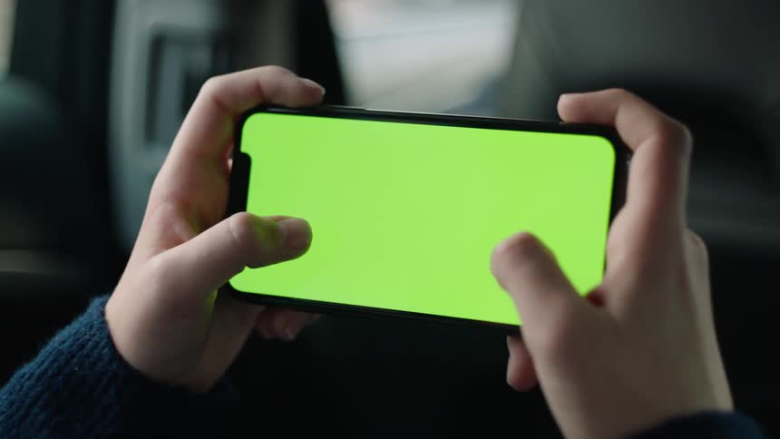Cinematic close-up of hands lifting a smartphone with a green screen and tracking markers in landscape mode inside a car during daylight. Perfect for VFX, screen replacement, app mockups, and projects about travel, mobility, or digital technology.